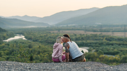 Fototapeta na wymiar Young man hugs his girlfriend on the mountain peak at sunset background.