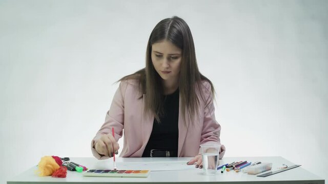 Attractive young woman draws with watercolors on a white paper at the table in a white studio