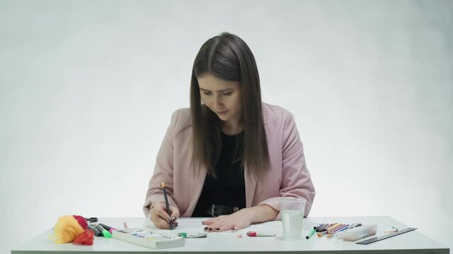 Attractive young woman draws with peencil on a white paper at the table in a white studio