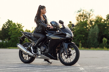 Stylish beautiful girl in black leather jacket and pants on outdoors parking sits on sports motorcycle and holds protective helmet.