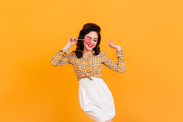 Shapely young woman in white skirt holding candy. Studio shot of pinup girl with lollipop dancing on yellow background.
