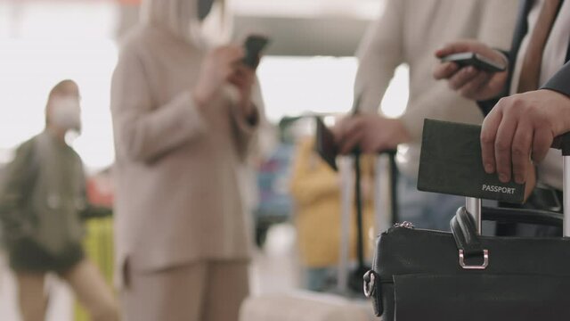 Slowmo closeup of unrecognizable businessman hand holding passport and luggage in selective focus while other people in protective masks standing in blurred background