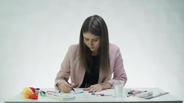 Attractive young woman draws with a black marker on a white paper at the table in a white studio