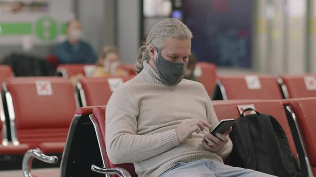 Medium close-up of senior caucasian man in protective mask using smartphone while waiting for flight in departure lounge during coronavirus