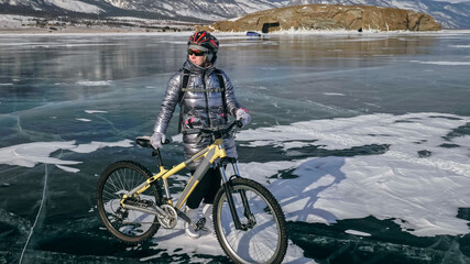 Woman stands near her bike on ice. The girl cyclist stopped to rest. The beautiful view of the sunset. The traveler is ride a cycle. Female is riding bike on the ice. Athlete puts on gear.