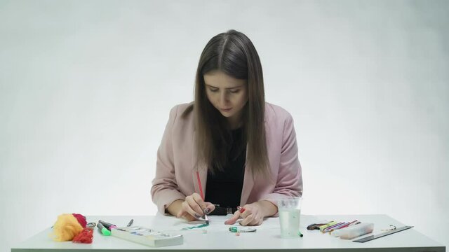 Attractive young woman draws with a red acrylic paints on a white paper at the table in a white studio