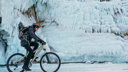 Man is riding bicycle near ice grotto. Rock with ice caves icicles. Teenage is dressed in black...