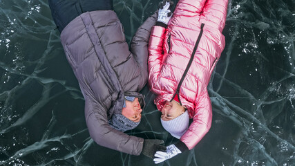 Couple has fun winter walk against background of ice of frozen lake. Lovers lie on clear ice with cracks have fun kiss and hug. View from above. Happy people on snow covered ice. Honeymoon love story. - obrazy, fototapety, plakaty