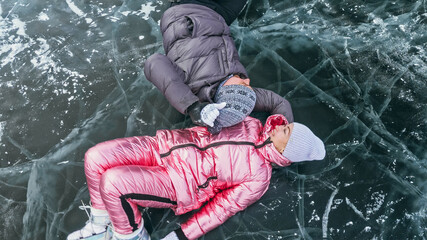 Couple has fun winter walk against background of ice of frozen lake. Lovers lie on clear ice with cracks have fun kiss and hug. View from above. Happy people on snow covered ice. Honeymoon love story.