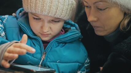 Obraz premium Attractive caucasian mother and daughter using smartphone sitting in winter cafe in jacket, hat. Advanced little child girl shows and explains to mom woman on smartphone, touching and watching.