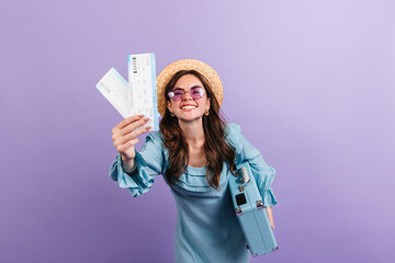 Full of life traveler in blue dress with funny facial expression shows plane ticket. Portrait of smiling girl in boater on lilac background
