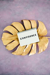Confidence notepad on top of golden leaf on pink background, psychology and attitude