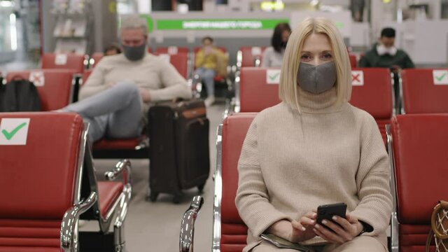 Panning slow-motion medium portrait of caucasian woman in protective mask looking at camera waiting for flight in departure lounge with other passengers