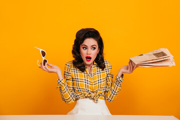Amazed pinup girl holding sunglasses on yellow background. Elegant young woman posing with newspaper.