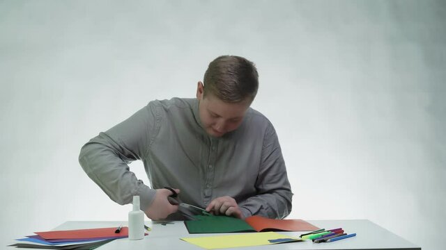 Young man uses scissors to cuts a green color paper at the table in a white studio