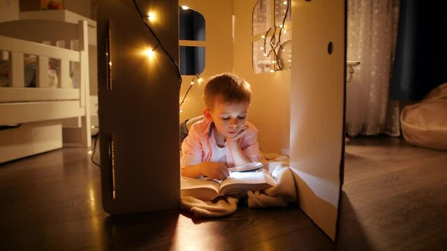 Panning shot of little smart boy reading big book at night with flashlight. Concept of child education and reading in dark room