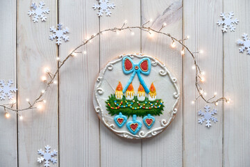 Gingerbread decorated with advent wreath with candles and hearts. Creative traditional gifts for children. Christmas flat lay with snowflakes, garland of lights. Rustic off white wooden table.