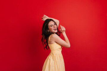 Mischievous curly lady is dancing on red background. Brunette in yellow dress sincerely smiles and enjoys photo shoot