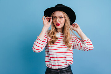 Curly woman in elegant hat looking at camera. Studio shot of fascinating blonde girl in striped shirt.