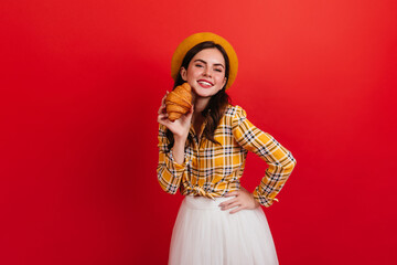 Parisian woman in stylish yellow shirt holding appetizing croissant. Lady in beret with smile looks into camera on red background