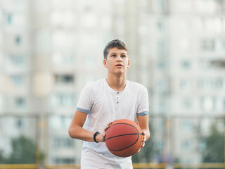 Cute young boy plays basketball on street playground in summer. Teenager in white t shirt with orange basketball ball outside. Hobby, active lifestyle, sport activity for kids.