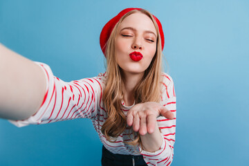 Romantic blonde girl sending air kiss. Studio shot of french lady taking selfie on blue background.