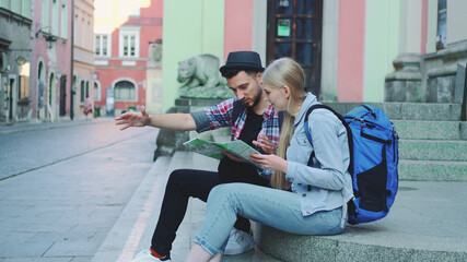 Happy tourist couple sitting on street stairs, checking city map and discussing. In the background there is beautiful old town view.