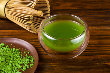 Bamboo matcha tea whisk also know as chasen with green matcha tea in a glass thermo cup and powdered matcha tea in a plate on a wooden background. 