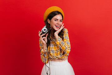 Dark-haired lady in orange beret is holding retro camera. Girl in bright blouse smiles coquettishly on red background