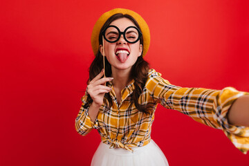 Lady in beret and round glasses shows tongue. Selfie of naughty girl in bright outfit on red background