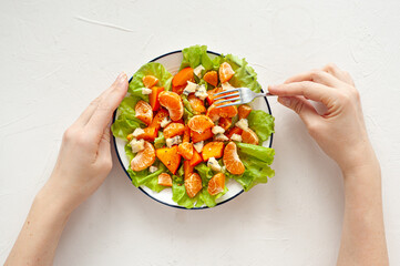 Bright vitamin salad with persimmon, tangerines and blue cheese on a white table. Women's hands hold a fork and a plate of salad. Dietary vitamin food. View from above.