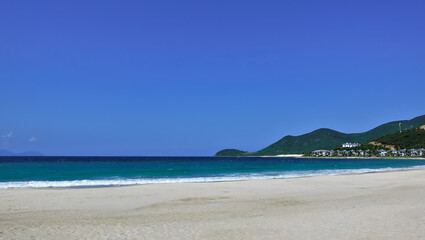 Turquoise waves foam on the white sand of the beach. A row of villas is visible in the distance. Green mountains against a clear blue sky. Vietnam. Nha Trang.