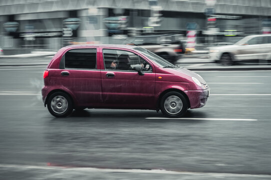 Compact city hatchback Daewoo Matiz with a woman driving. Fast moving magenta car on the city wet road. Overspeed concept.