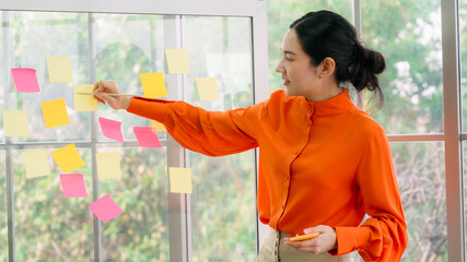 Business people work on project planning board in office and having conversation with coworker friend to analyze project development . They use sticky notes posted on glass wall to make it organized .