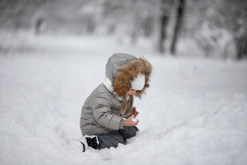 little girl in a gray jacket with fur in winter sits on the snow and makes a snowball 
outdoor snow games