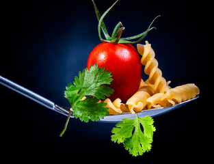 Tomato in a spoon with fresh greens and pasta on a dark black background. Tomato in a spoon close up
