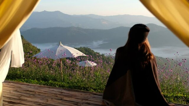 Rear view of a woman sitting on wooden balcony while watching sunrise and a beautiful mountains view outside the tent