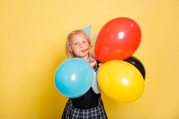 Fototapeta na wymiar A girl in a school uniform and a festive cone on her head holds balloons in her hands and blows a pipe isolated on a yellow background.