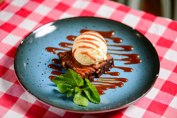 Traditional recipe brownie with ice cream served on a blue plate over table with red plaid tablecloth.