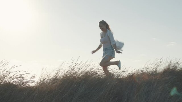 Blonde woman walking and running in tall grass. 50 fps gimbal moving shot, wind blowing her hair and white shirt in sunset