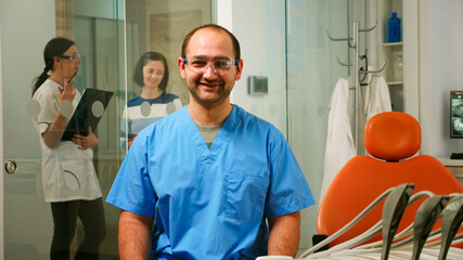 Portrait of smiling man nurse in dental office while pediatric doctor is talking with patient in background. Stomatologist assistant looking at camera sitting on chair in stomatological clinic.