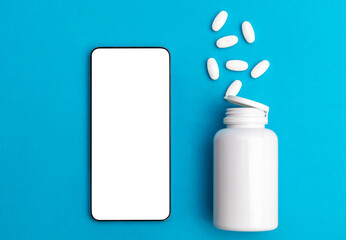 Smartphone with blank screen and plastic medical bottle with scattered pills on blue. Copy space.