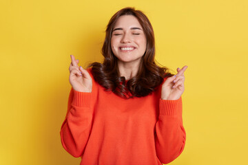 Portrait of overjoyed young caucasian woman with dark hair lifting fists with crossed fingers from joy or happiness, keeps eyes closed and clenching teeth, hopes for success.