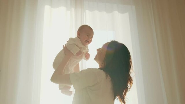 Young happy mother holds an adorable infant in her hands and lifts her up. Mom plays with daughter in the bedroom 