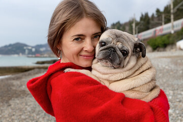A pug in the arms of a woman in a red coat. Warm winter day by the sea.