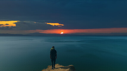 The man with a backpack standing on a mountain against a sea sunrise background