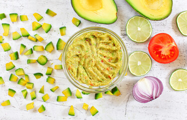 Avocado halves and pieces, limes tomato, onion - basic guacamole ingredients on white working board, finished mix in small glass bowl near