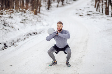 Bodybuilder lifting kettlebell while crouching in woods on snowy trail at winter. Bodybuilding, winter sport, fitness
