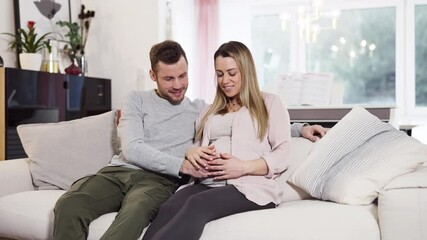 Young pregnant happy couple sitting on couch husband stroking baby belly of blond wife shot in 4k - Powered by Adobe