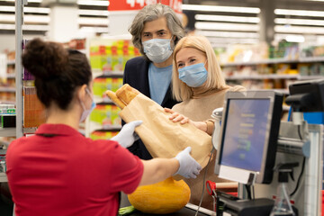 Mature man and woman in protective masks passing packet with bread to cashier